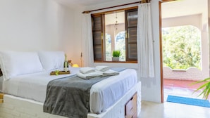 A neatly made bed with white linens, a grey blanket, and two folded towels. There is a small table with a bottle and glasses on it. A window with wooden shutters and a potted plant is visible. - Sitio Natureza piscina e churrasqueira (Ibiuna)