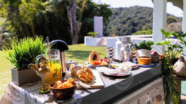 An outdoor dining setup with a variety of food items, a pitcher of juice, and a decorative rooster figurine. - Sitio Natureza piscina e churrasqueira (Ibiuna)