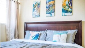 Une chambre à coucher avec une tête de lit en bois, un lit recouvert d’une couette grise et blanche, et trois tableaux abstraits accrochés au mur.