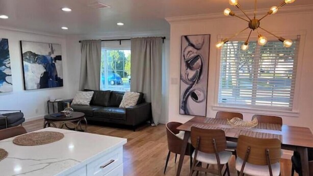 A modern living room with a sofa, dining table, and chairs. There is a large window with curtains, artwork on the walls, and a ceiling light fixture. - Buena Park Modern Home/Knott's/Disney/Beach

 (Buena Park)