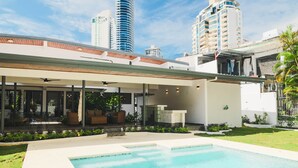 Un espace piscine extérieur moderne, doté d’un coin salon couvert, entouré de verdure et avec de grands bâtiments en arrière-plan.