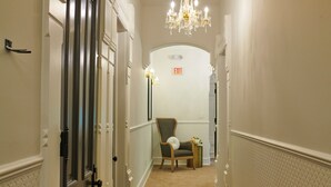 A hallway with a chandelier, an exit sign, a chair, and a trash can. - Mon Bijou (Oconomowoc)