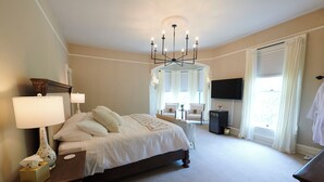 A bedroom with a large bed, bedside lamps, a TV, and a window with curtains. - Mon Bijou (Oconomowoc)