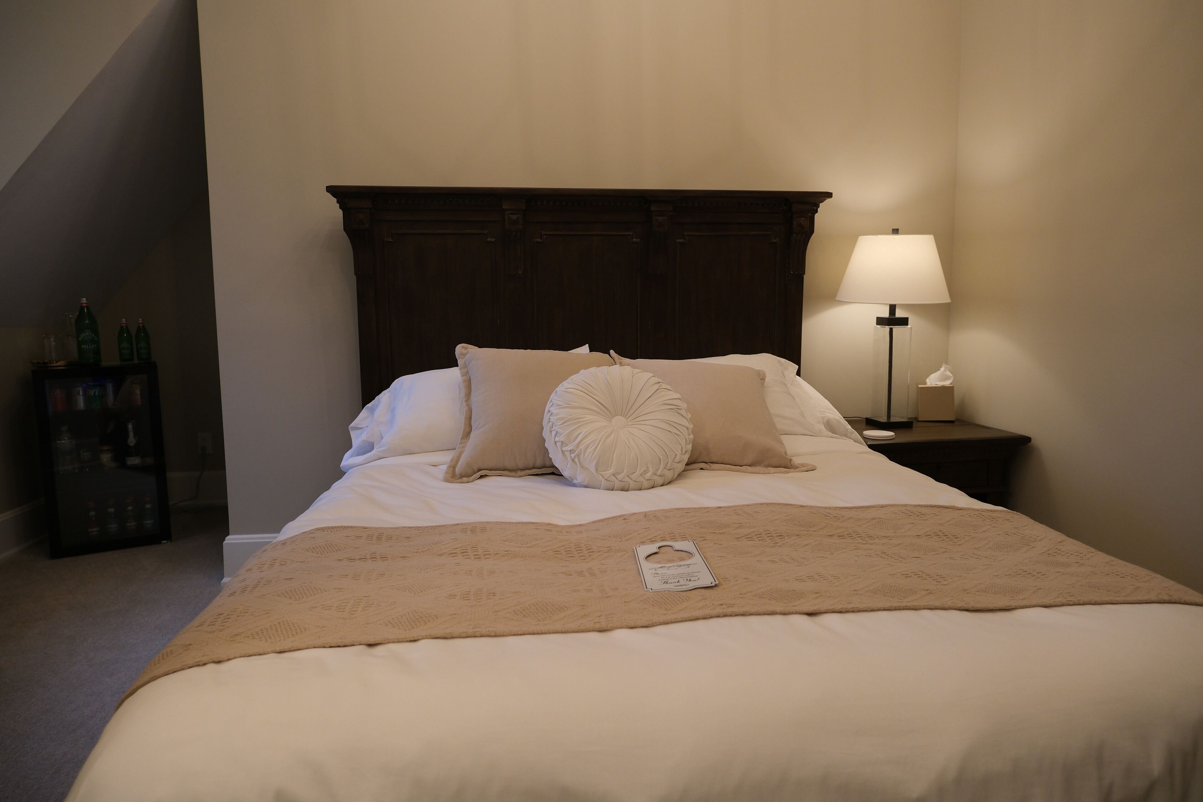 Una cama bien hecha con un edredón color crema, dos almohadas y una etiqueta en el edredón. Se ve una mesita de noche con una lámpara y una botella de agua. Se observa un cabecero de madera y una pared con una estantería donde hay botellas.