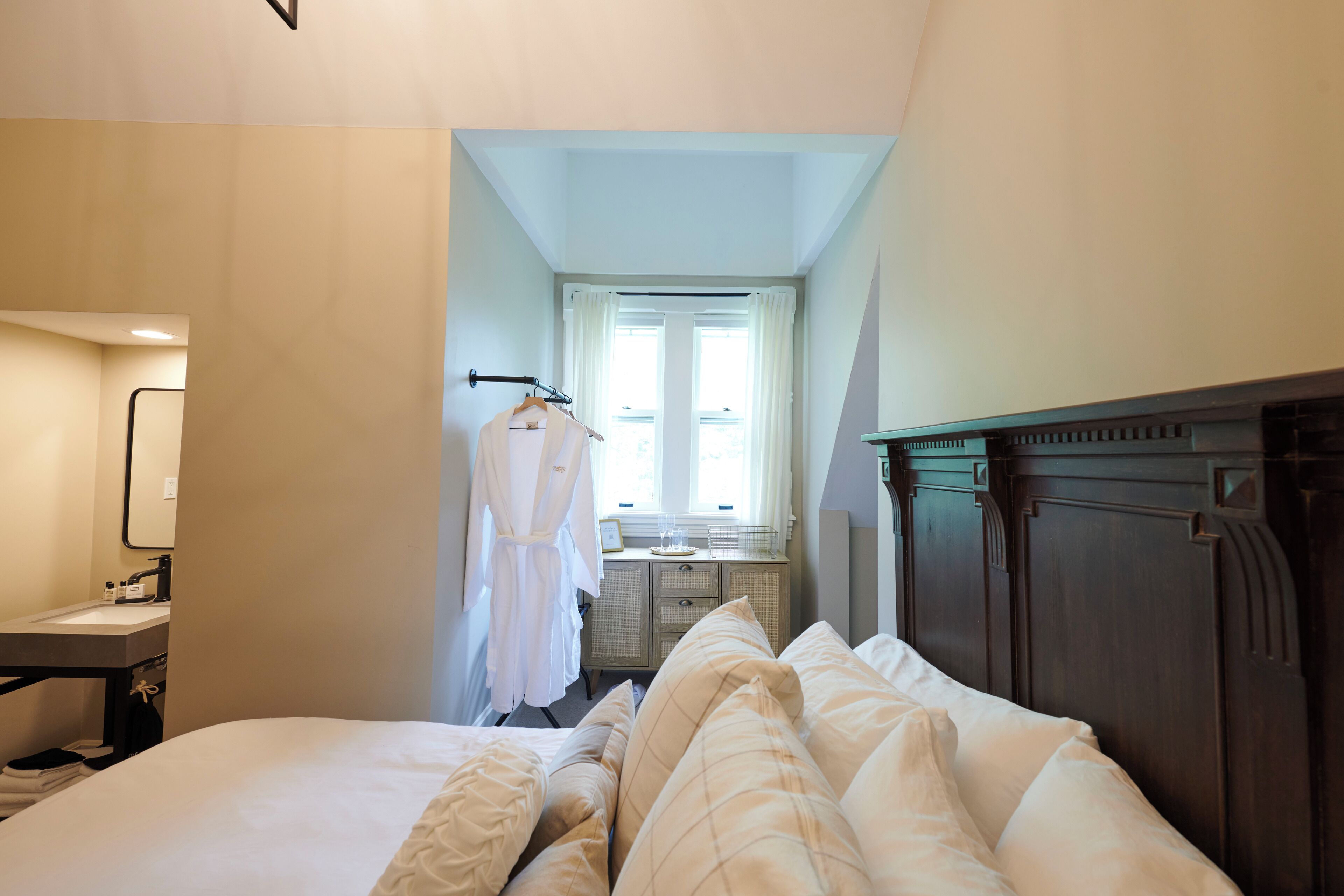 Habitación de hotel con cama, tocador con espejo, bata colgada en la pared y ventana con cortinas.