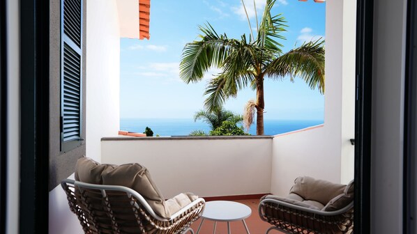 A balcony with two wicker chairs and a small round table, overlooking a palm tree and the sea.