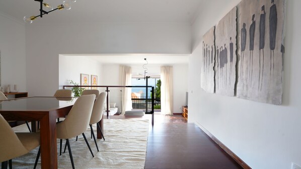 A spacious dining area with a long table and chairs, a large abstract painting on the wall, and a view through the glass door to the outside.