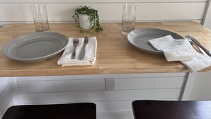 Uma mesa de jantar bem posta com dois pratos, talheres, copos, uma pequena planta em vaso e um guardanapo branco sobre uma mesa de madeira.