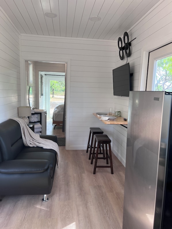 A compact kitchen with a built-in refrigerator, wooden stools, a dining table, and a TV mounted on the wall.