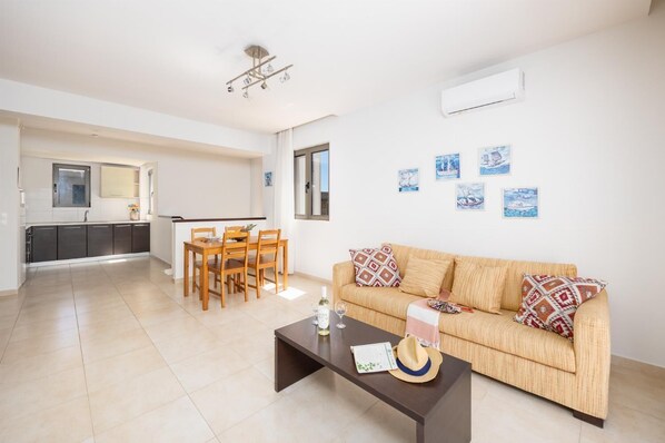 A modern living room with a sofa, coffee table, and dining area. There is a kitchen in the background with a window and an air conditioning unit.
