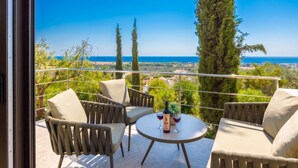 Ein Balkon mit Sitzgelegenheit im Freien, einem Tisch mit Flasche und Gläsern sowie Blick auf das Meer und die Landschaft.