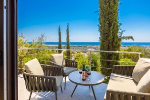 Ein Balkon mit Sitzgelegenheit im Freien, einem Tisch mit Flasche und Gläsern sowie Blick auf das Meer und die Landschaft.