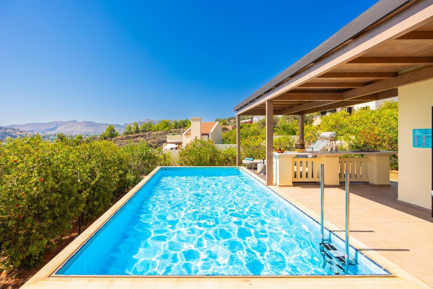 A rectangular swimming pool with clear blue water, surrounded by a covered patio area with lounge chairs and a barbecue grill.