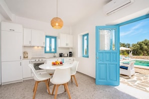 A modern kitchen with white cabinets, a dining table with chairs, a fruit bowl, and a blue door leading to a pool area.