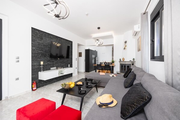 A modern living room with a grey sofa, red ottoman, and a black coffee table. A flat-screen TV is mounted on a black brick wall. - Villa Elysian - Two Bedroom Villa, Sleeps 4 (Afandou)