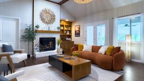 A living room with a fireplace, a sofa, a coffee table, and a large window. - Serenity WR Deluxe Escape Indoor Pool (Warner Robins)