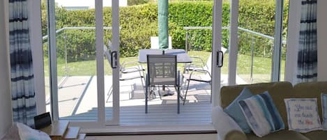 Ein gemütliches Wohnzimmer mit einer Couch und einem Tisch mit Stühlen, eine Glastür, die auf eine Terrasse im Freien mit Tisch und Stühlen führt.