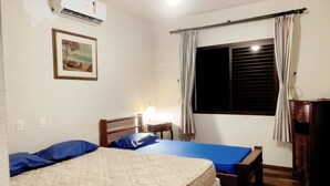 A bedroom with two beds, a nightstand, a lamp, a window with curtains, and an air conditioning unit.