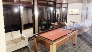 A pool table in a room with a tiled floor, a sofa, and a glass partition with a view of palm trees.