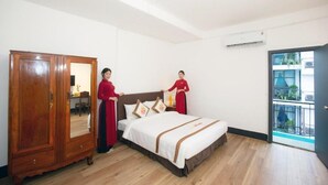 Une chambre d’hôtel avec une armoire en bois, un lit recouvert d’une courtepointe blanche et marron, et une vue sur un balcon par une porte ouverte.