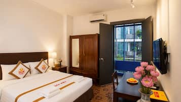 A hotel room with a bed, bedside table, lamp, wardrobe, and a desk with a vase of flowers.