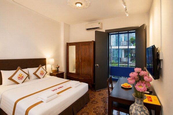 A hotel room with a bed, bedside table, lamp, wardrobe, and a desk with a vase of flowers. - The Rice Hotel & Tour - 131 Bui Vien (Ho Chi Minh City)