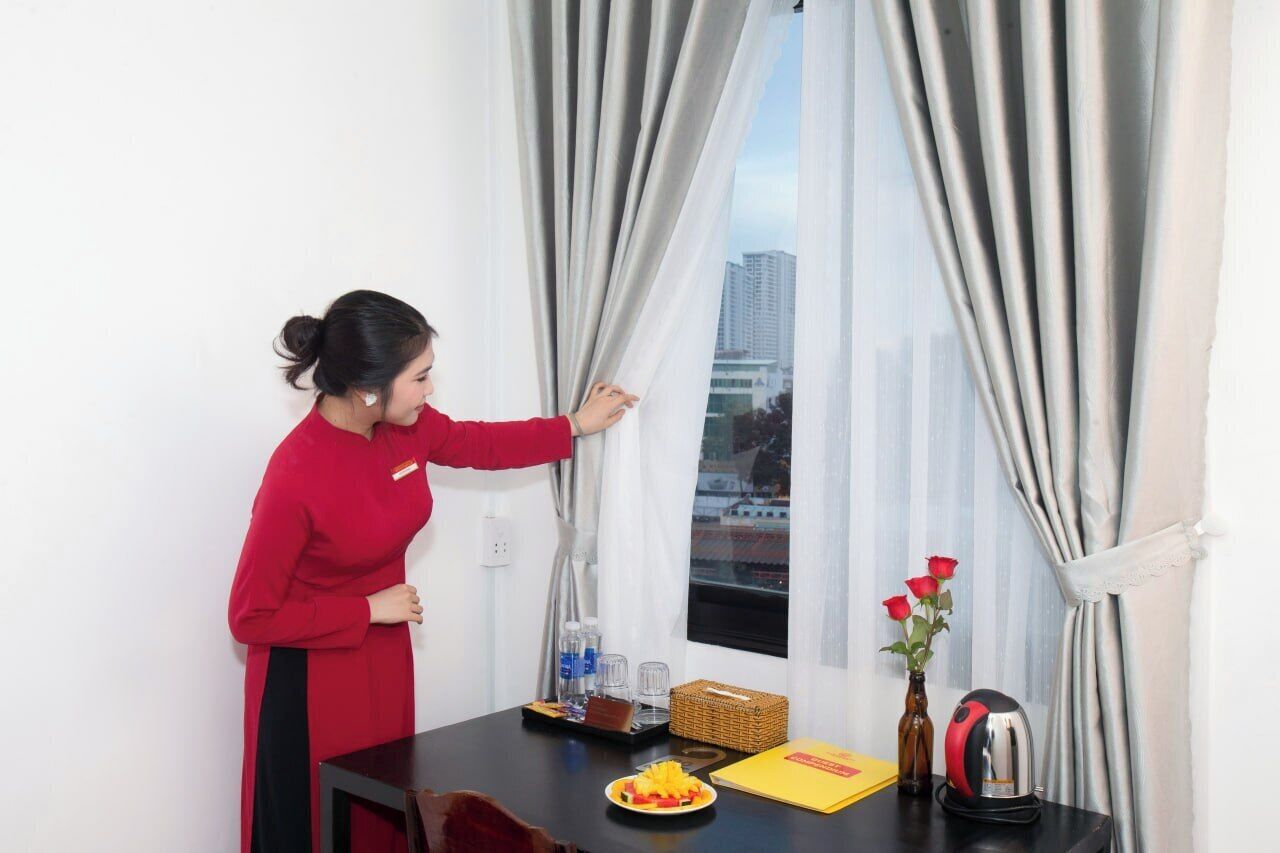 A person in a red uniform adjusting curtains in a room with a table that has a plate of food, a bottle, a basket, and a kettle.