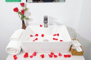 Un baño con un lavamanos blanco, un florero con rosas rojas, una toalla enrollada y una cesta de artículos de tocador.