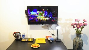 A hotel room with a TV displaying a cityscape, a vase with pink flowers, a coffee maker, a plate of fruit, and a "NO Smoking" sign. - The Rice Hotel & Tour - 131 Bui Vien (Ho Chi Minh City)