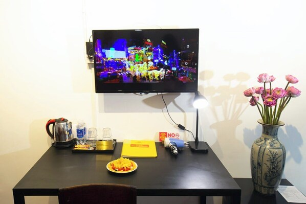 Habitación de hotel con un televisor mostrando un paisaje urbano, un florero con flores rosas, una cafetera, un plato de fruta y un cartel que indica 
