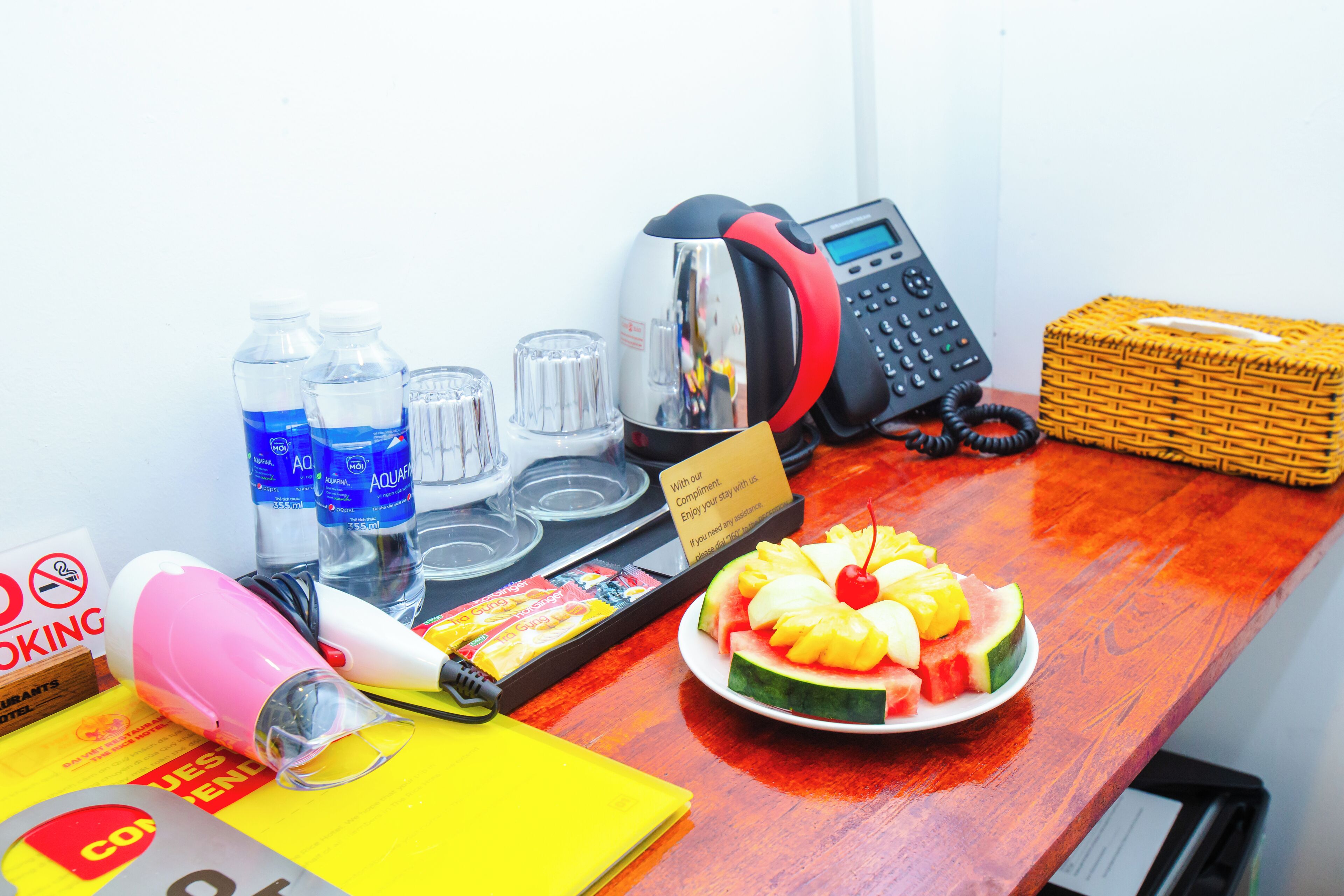 A desk with a phone, water bottles, a hairdryer, a plate of fruit, and a basket.