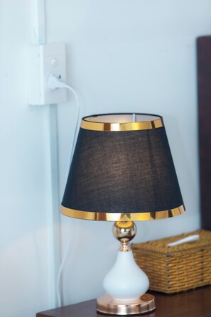 Una lámpara de mesa con base blanca y pantalla negra con detalles dorados, conectada a un enchufe de pared.