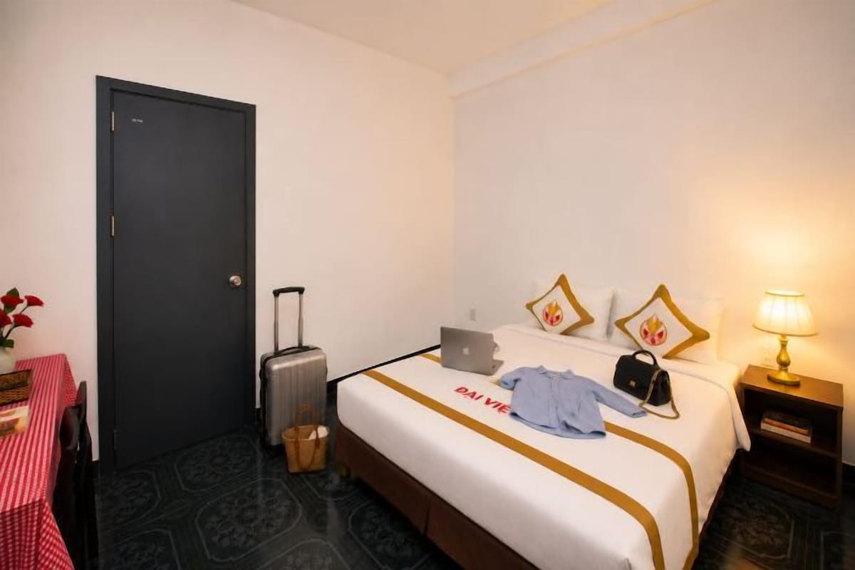 A hotel room with a bed, a desk, a chair, a suitcase, a laptop, and a lamp.