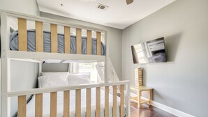 Uma cama beliche infantil, com estrutura de madeira e roupa de cama branca, uma pequena mesa de madeira e uma televisão de tela plana fixada na parede.
