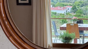 Een kamer met een spiegel met houten lijst, die een balkon weerkaatst met een tafel, stoelen en uitzicht op een heuvel met huizen.