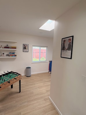 Ett rum med ett fotbollsspel, ett fönster med persienner, en inramad affisch på väggen och en hylla med böcker.