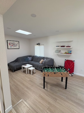 Ett vardagsrum med en soffa, ett soffbord, ett fotbollsspel, en projektor och hyllor med böcker och leksaker.