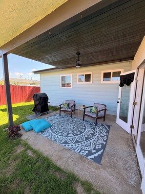 A covered patio area with two chairs and a patterned rug, a ceiling fan, and a sliding glass door.