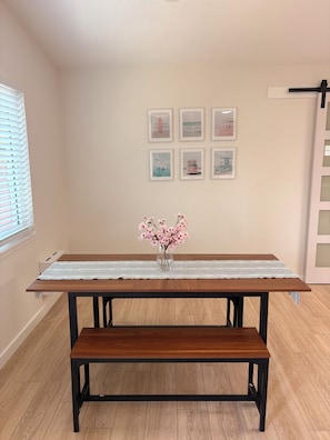 A room with a wooden table, bench, and a vase with pink flowers. There are six framed pictures on the wall.