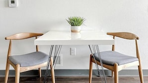 A dining area with a white table, two wooden chairs with grey cushions, a small potted plant, and a wall-mounted artwork. - Sleepover | Lovely 1BD/BA + High Ceilings - DSM (West Des Moines)