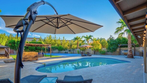 A swimming pool with lounge chairs and an umbrella, surrounded by palm trees and a covered patio area. - Backyard Paradise in Chandler (Chandler)