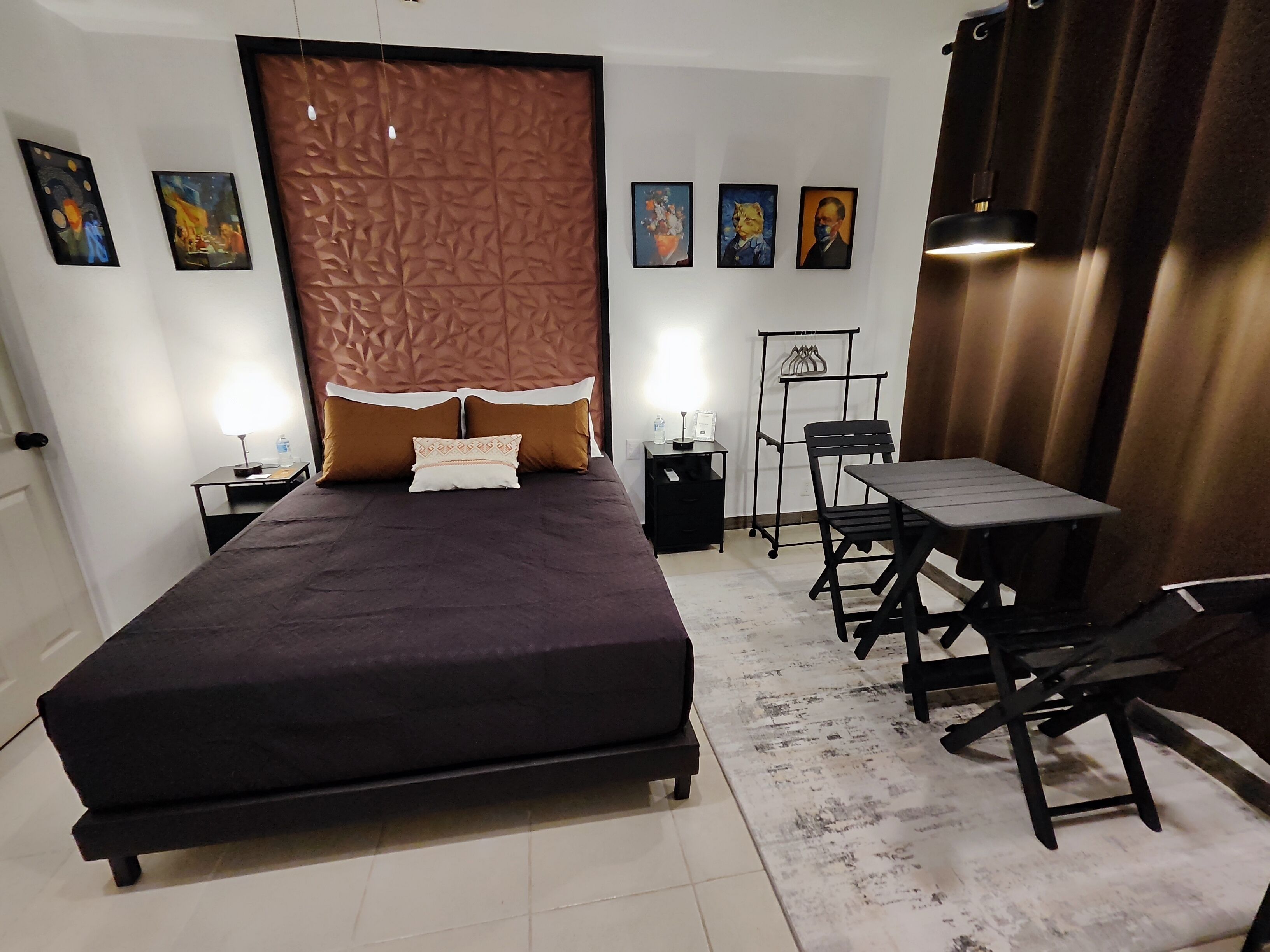 A bedroom with a bed, bedside tables, a desk, and chairs. There are wall-mounted artworks and a curtain.