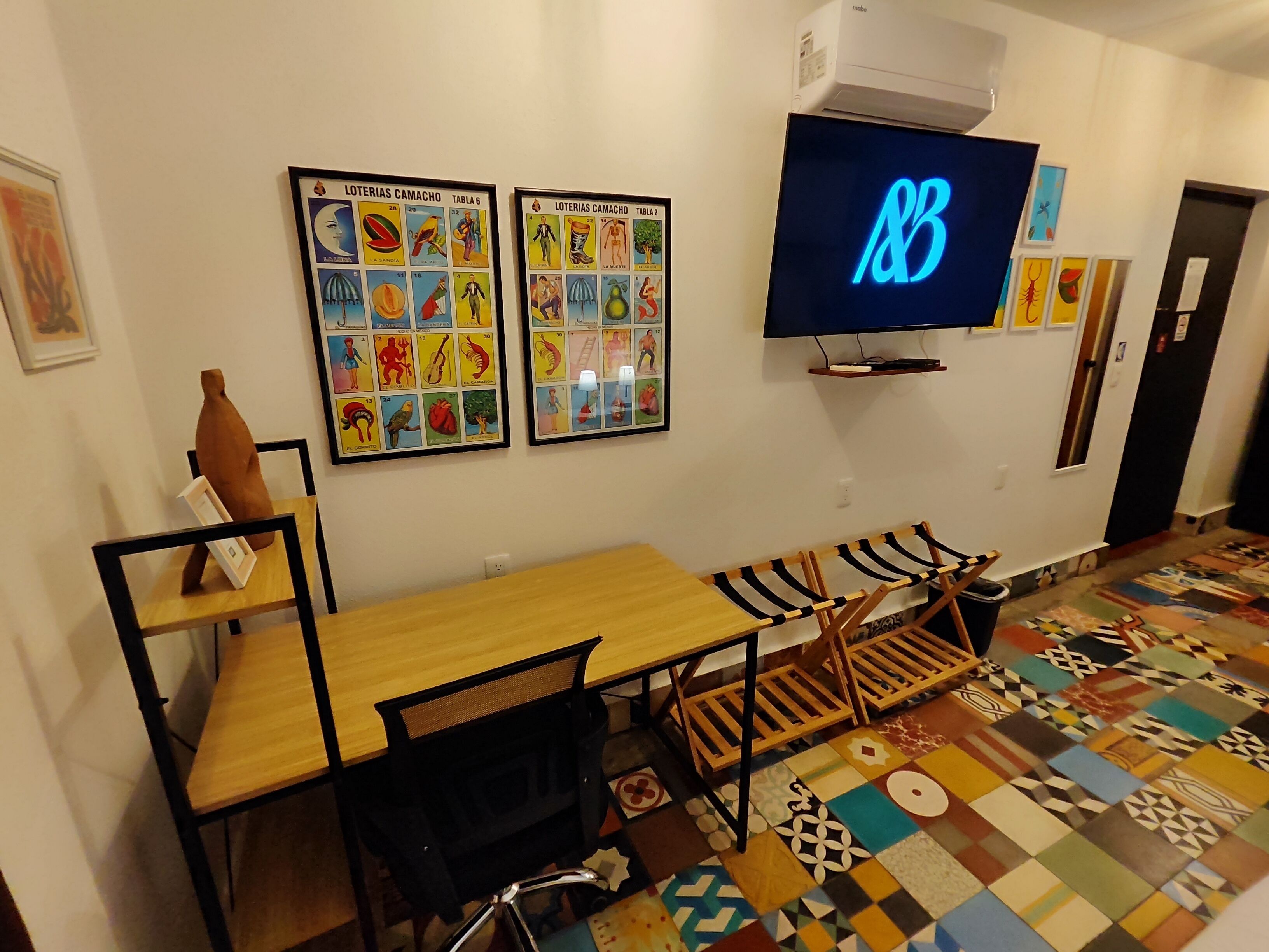 A room with a wooden table, a black chair, a TV mounted on the wall, and a colorful patterned floor.