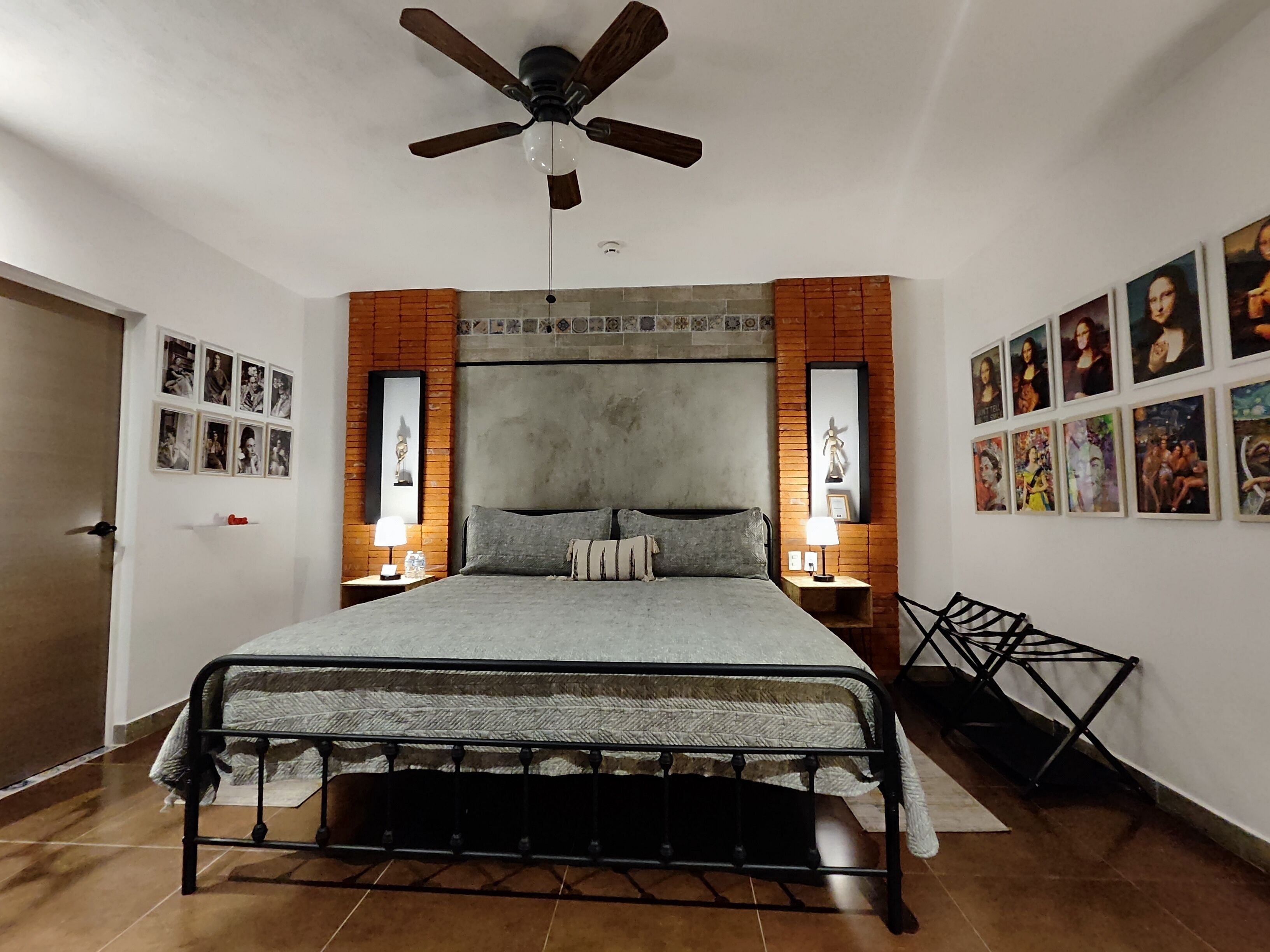 A bedroom with a bed, two bedside tables with lamps, a ceiling fan, and a brick accent wall.