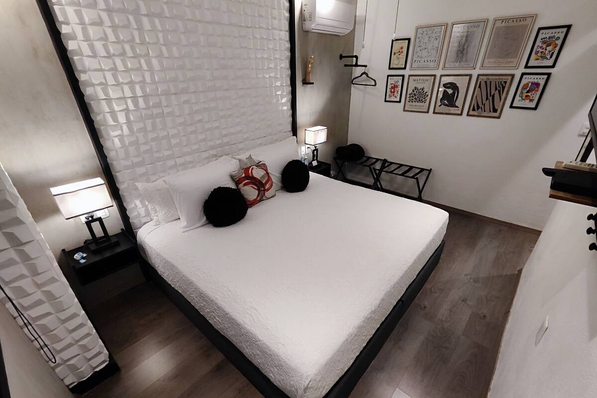 A modern bedroom with a large bed, a nightstand with a lamp, a chair, and artwork on the wall.