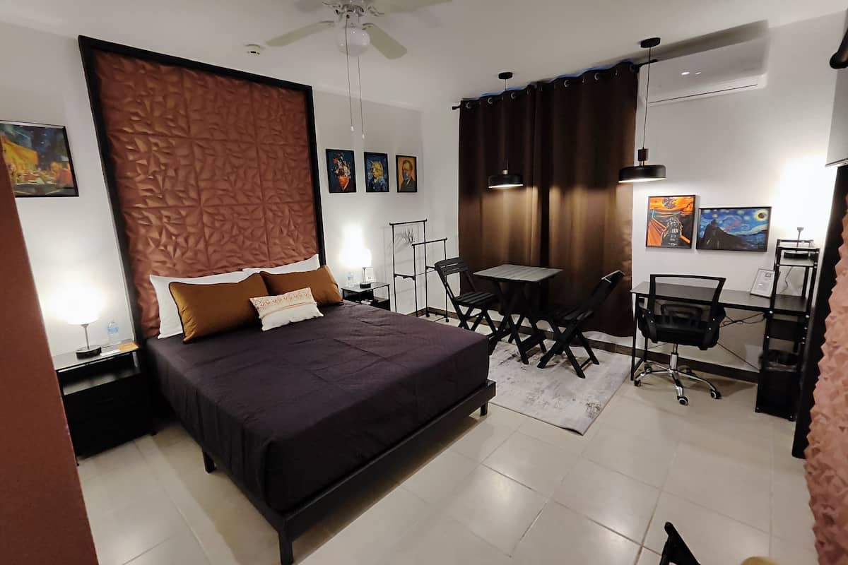 A modern hotel room with a large bed, a desk, and a chair. There is a ceiling fan, a window with curtains, and artwork on the walls.