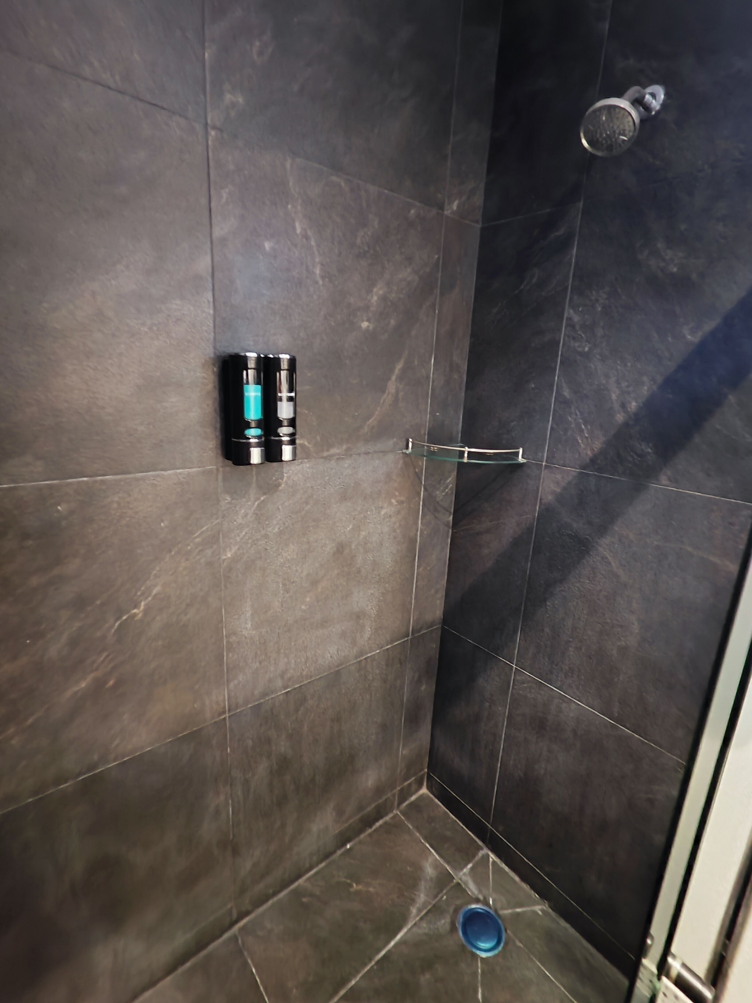 A bathroom with dark tiles, a shower, a glass door, and a blue bowl on the floor.