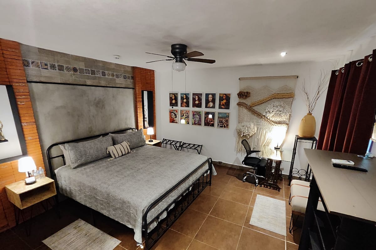 A bedroom with a bed, a desk, a chair, a ceiling fan, and artwork on the wall.