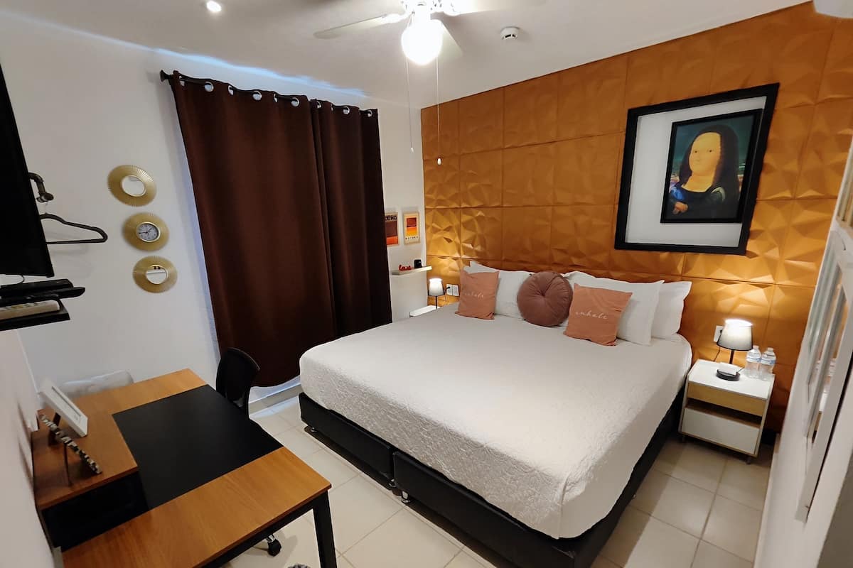 A modern hotel room with a large bed, a desk, a chair, a trash bin, a nightstand with a lamp, and a painting on the wall.