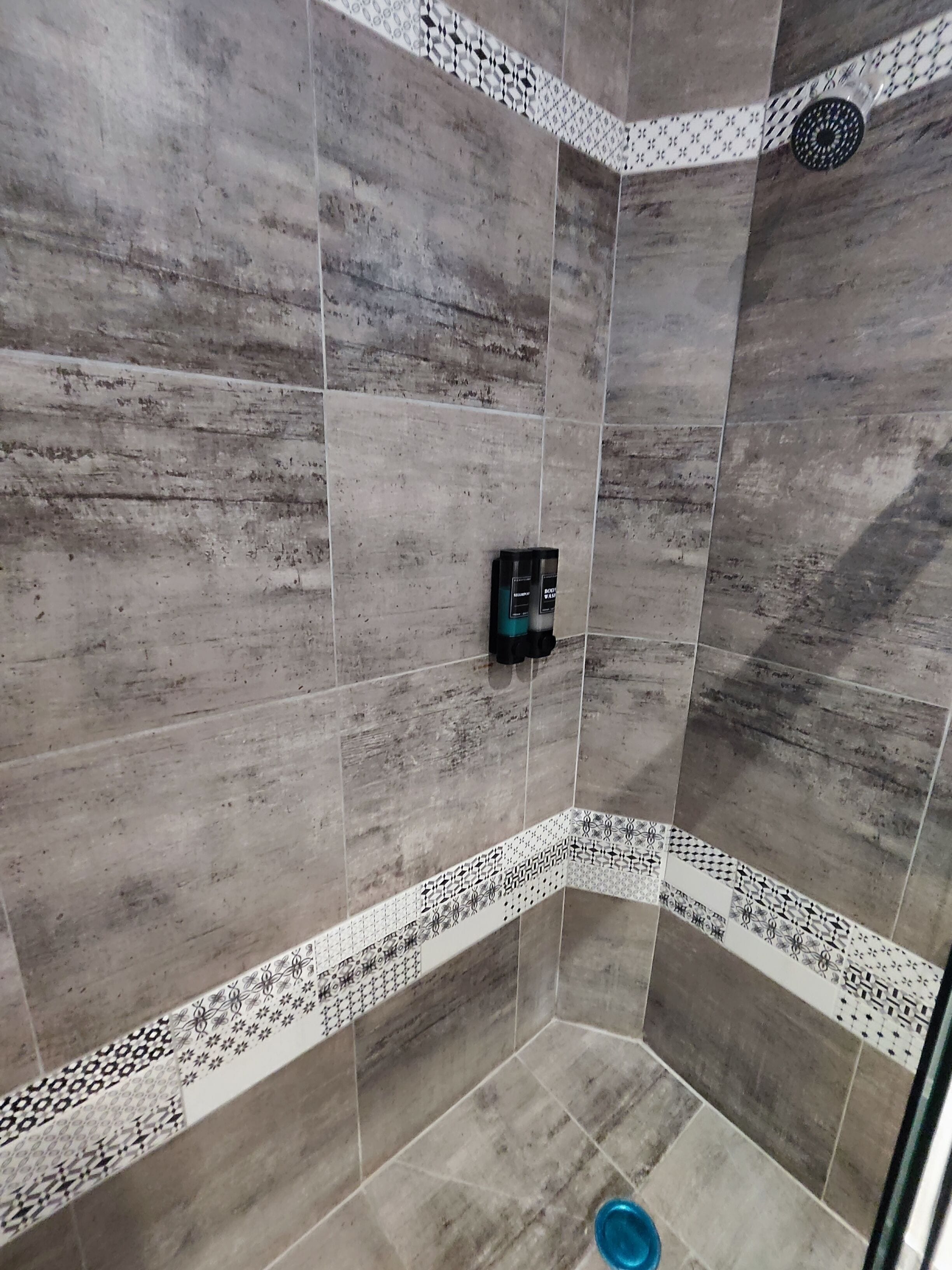 A shower with grey tiles, a black soap dispenser, and a blue basin.